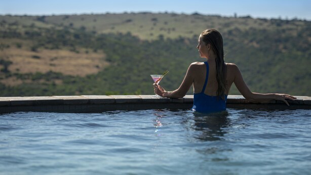 A person in a blue swimsuit sitting by a pool with a drink in hand, overlooking a hilly landscape.