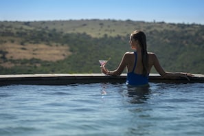 A person in a blue swimsuit sitting by a pool with a drink in hand, overlooking a hilly landscape. - Founders Lodge by Mantis (Paterson)