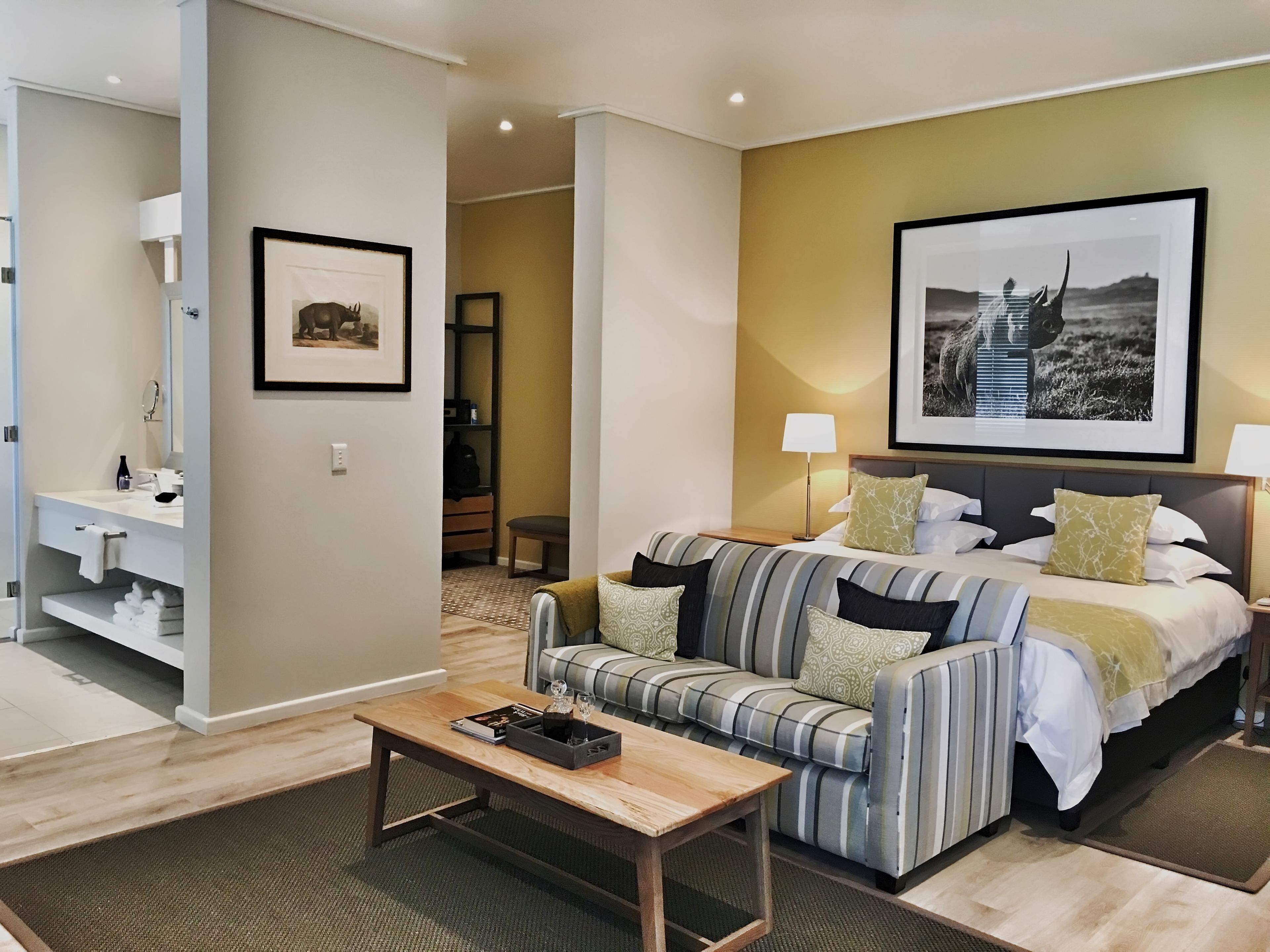 A hotel room with a bed, a sofa, a bedside table, a lamp, a framed picture on the wall, and a bathroom with a sink and towel rack.