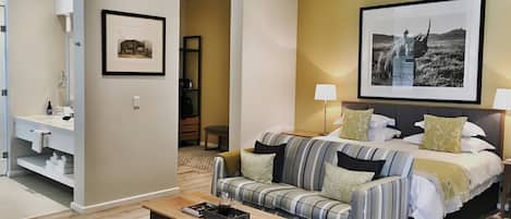 A hotel room with a bed, a sofa, a bedside table, a lamp, a framed picture on the wall, and a bathroom with a sink and towel rack.