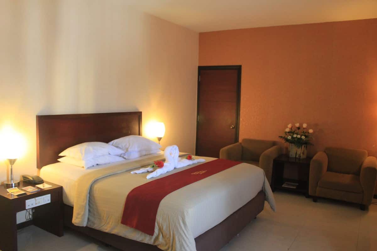 Habitación de hotel con cama, lámparas de noche, una silla, mesita de noche y un florero con flores.