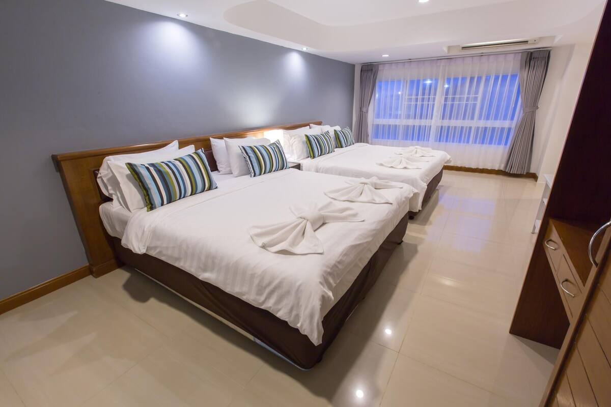 A hotel room with two beds, a wooden headboard, a large window with curtains, and a tiled floor.