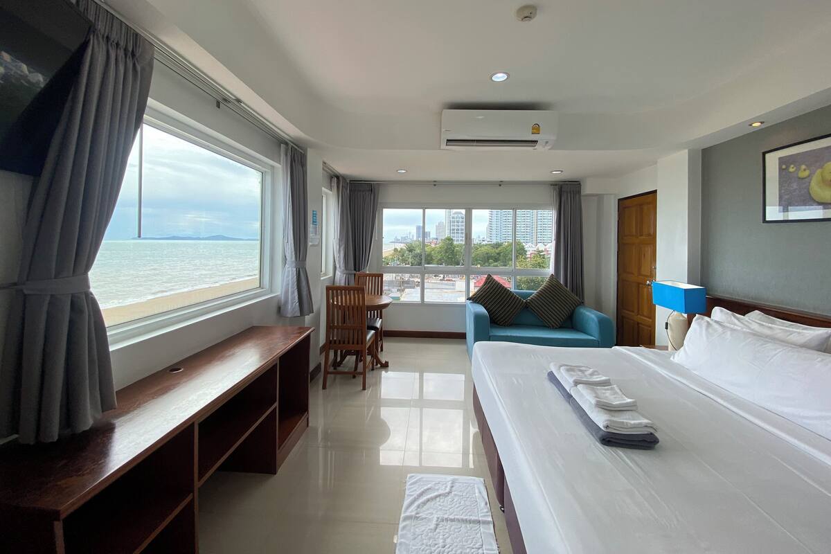 A hotel room with a bed, a desk, a chair, a blue sofa, and a view of the sea and cityscape.