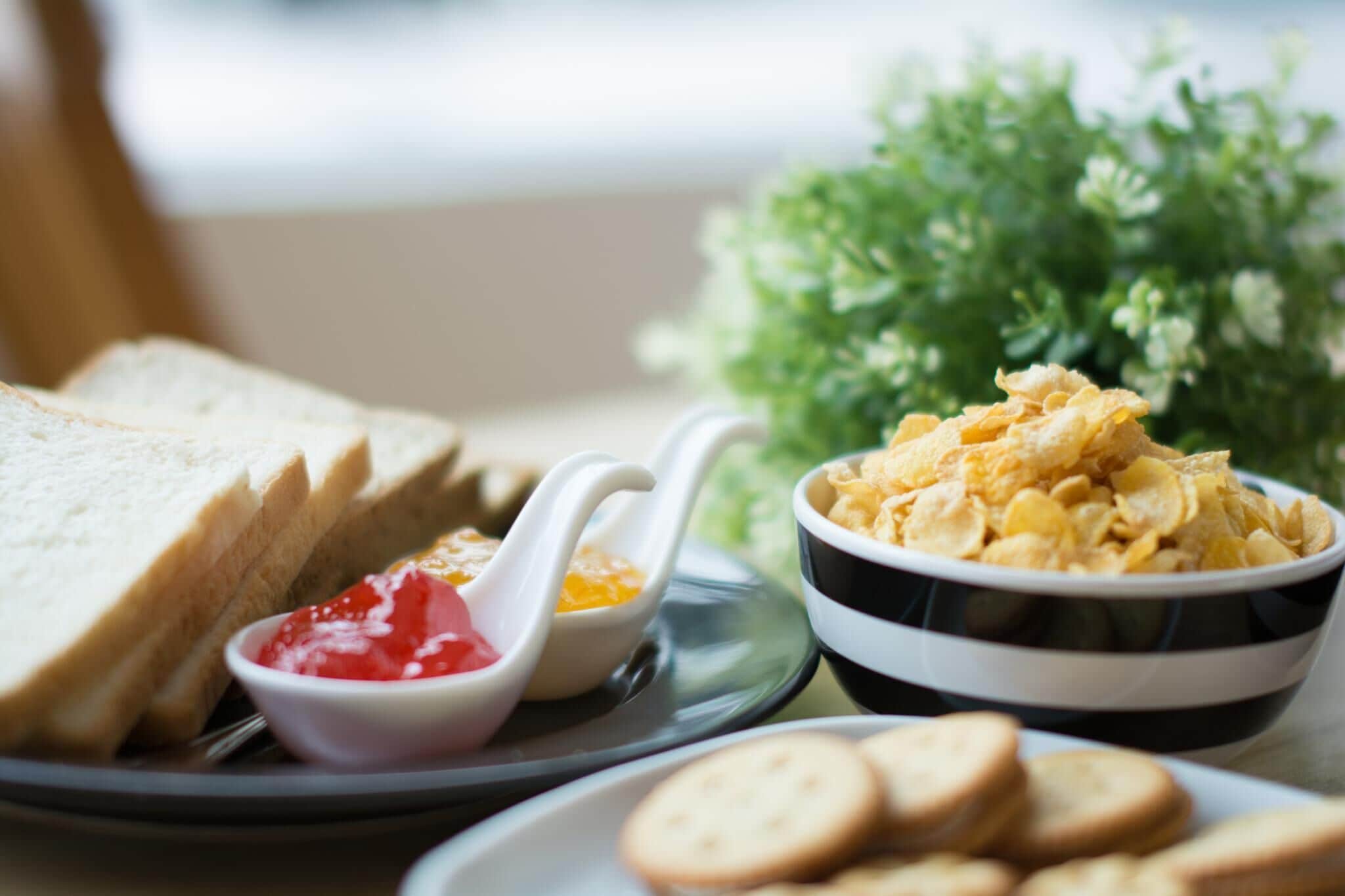 A plate with slices of white bread, a bowl of cereal flakes, a small bowl of jam, and a few biscuits.