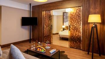 A modern hotel room with a wooden floor, a sofa, a coffee table with fruit and snacks, a TV, and a bed with a nightstand.