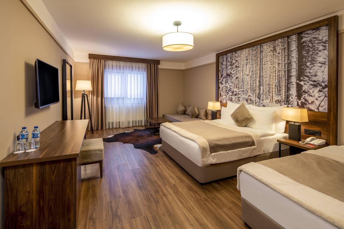 Une chambre d’hôtel avec un plancher en bois, un grand lit, un bureau avec un téléviseur à écran plat, un canapé, une grande fenêtre avec des rideaux et un téléphone fixé au mur.