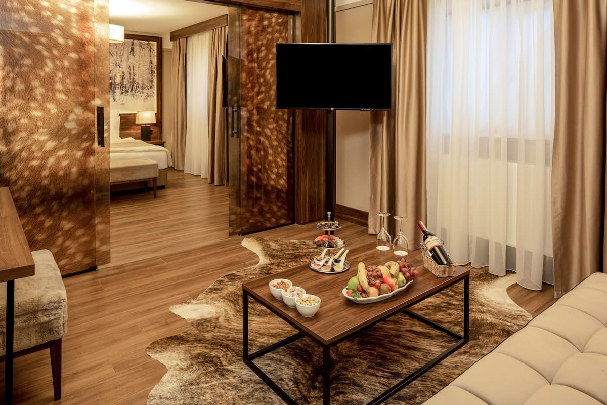 Une chambre d’hôtel moderne avec un plancher en bois, une grande télévision à écran plat, une table à manger avec des fruits et des en-cas, un canapé et une grande fenêtre avec des rideaux.