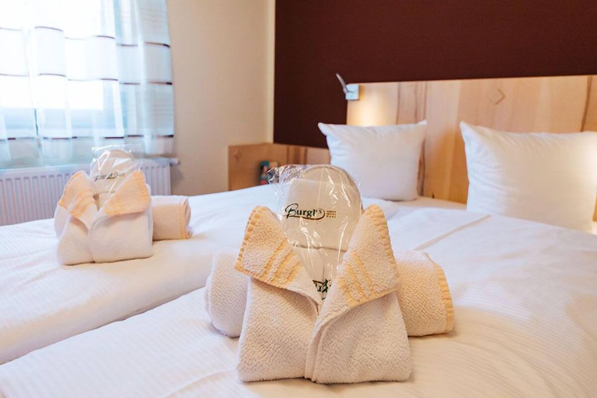 A hotel room with neatly folded towels on the bed, a wooden headboard, and a window with blinds.