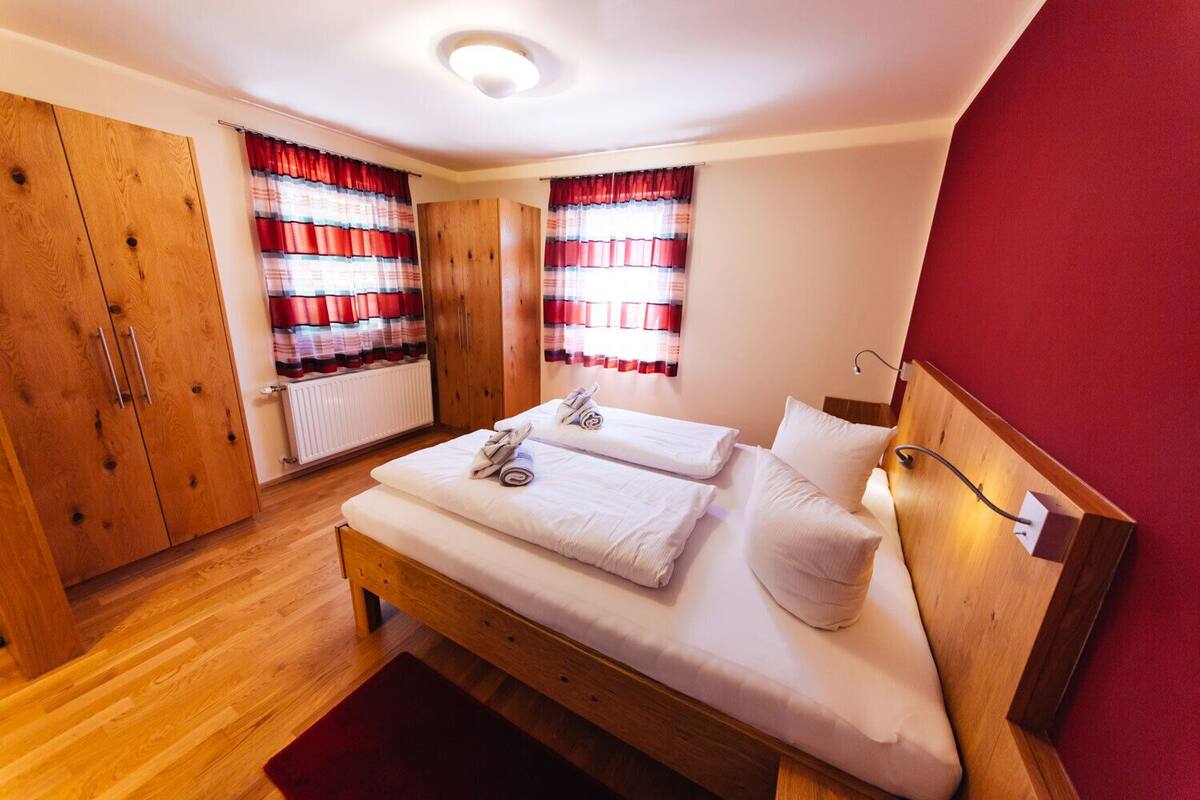 A bedroom with a wooden bed, two pillows, a red wall, and a window with red curtains.