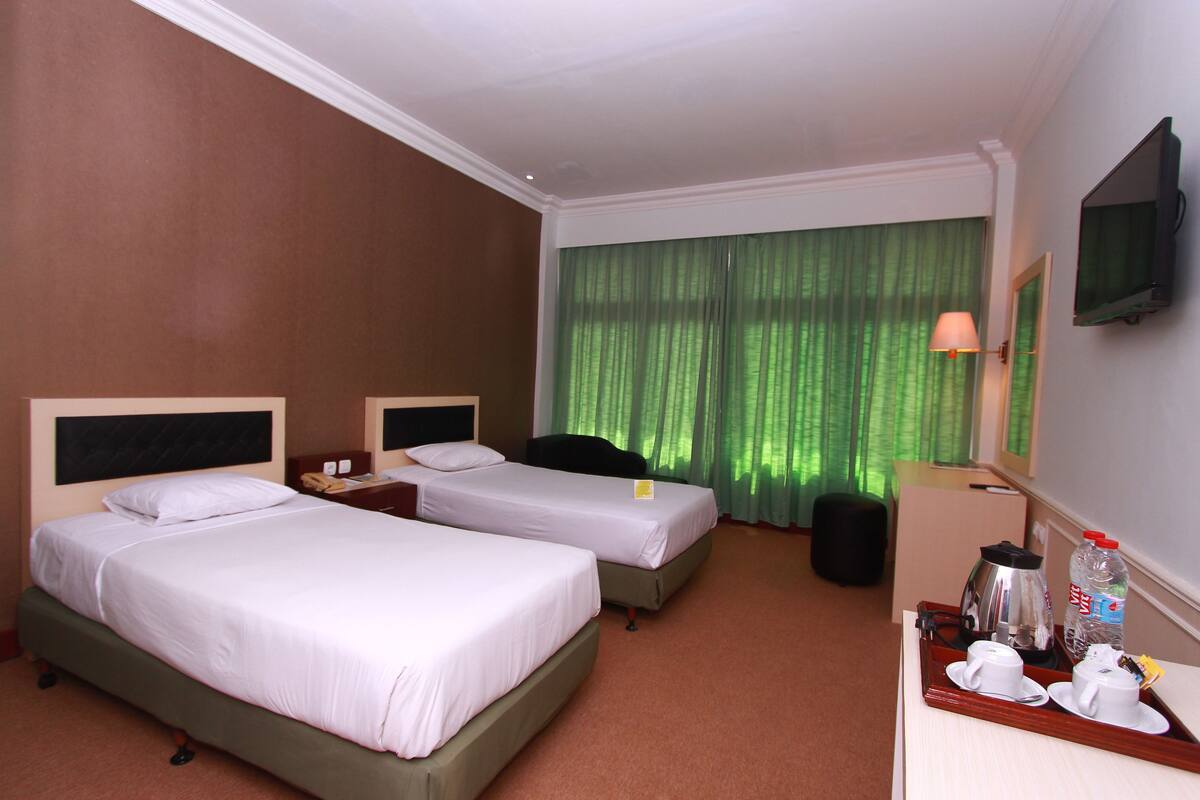 Habitación de hotel con dos camas individuales, un escritorio con silla, un televisor montado en la pared y una ventana con cortinas verdes.