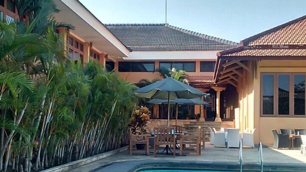 A swimming pool with lounge chairs and umbrellas, surrounded by palm trees and a building with a tiled roof. - Mutiara Hotel Cilacap (Cilacap)