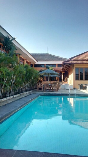 A swimming pool with lounge chairs and umbrellas, surrounded by palm trees and a building with a tiled roof. - Mutiara Hotel Cilacap (Cilacap)