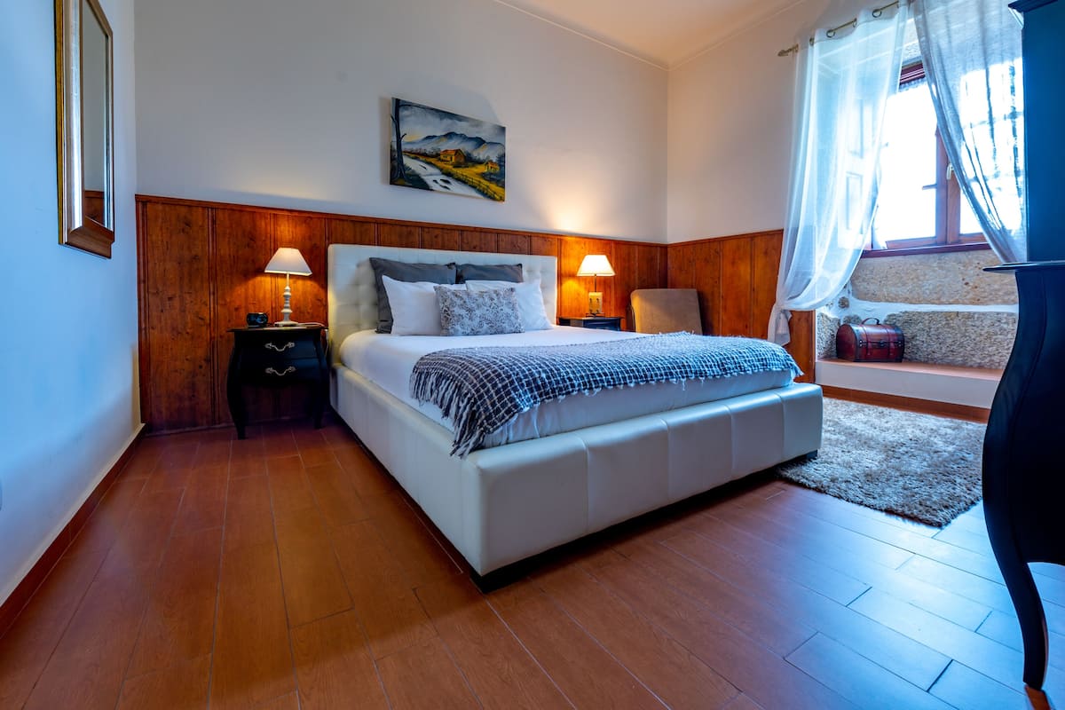 Ein Schlafzimmer mit einem großen Bett, Holzwänden, zwei Nachttischlampen, einem Gemälde an der Wand und einem Fenster mit durchsichtigen Vorhängen.