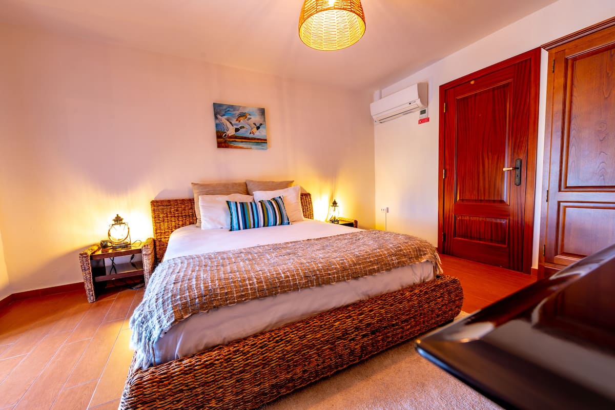 Ein Schlafzimmer mit einem großen Bett, einem Holzschrank, einem Bild an der Wand und einem Korbstuhl.