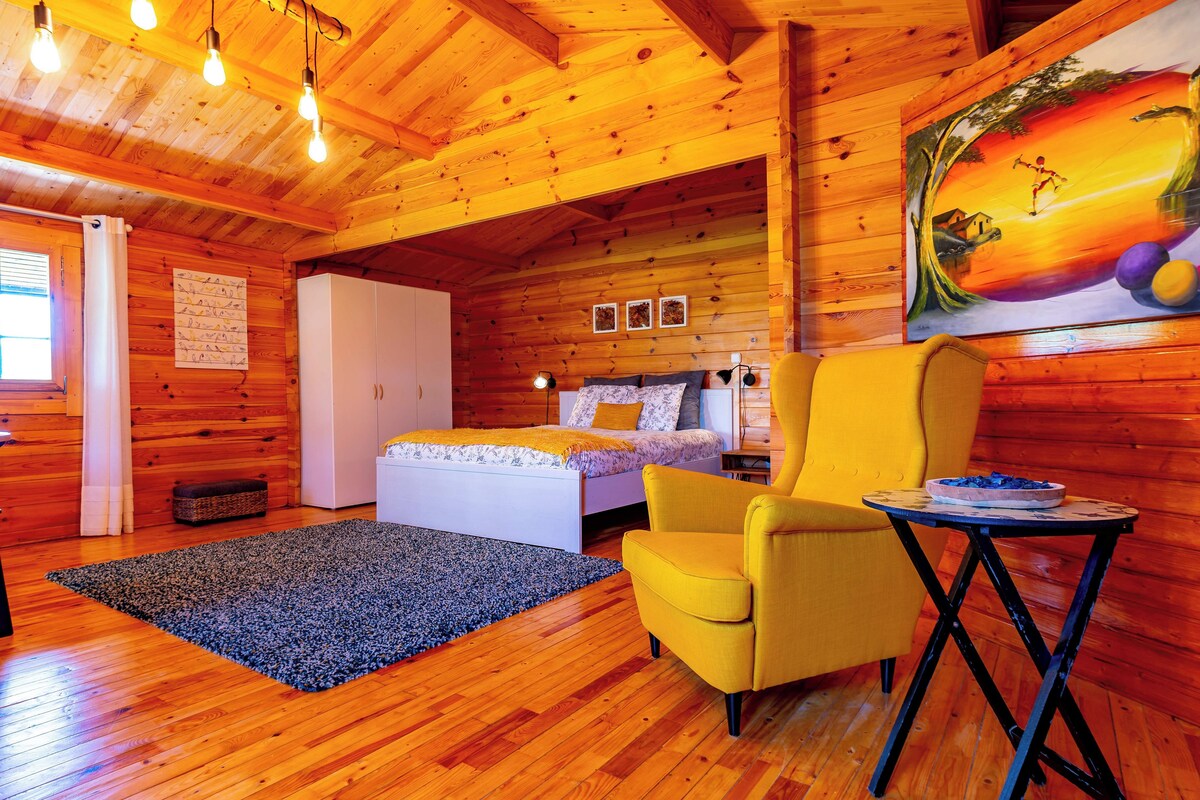 A wooden cabin interior with a bed, a yellow armchair, a small table, and a painting on the wall.