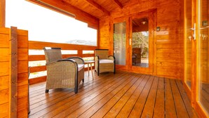 A wooden cabin with a wooden deck, wicker chairs, and a glass door leading to an outdoor area.