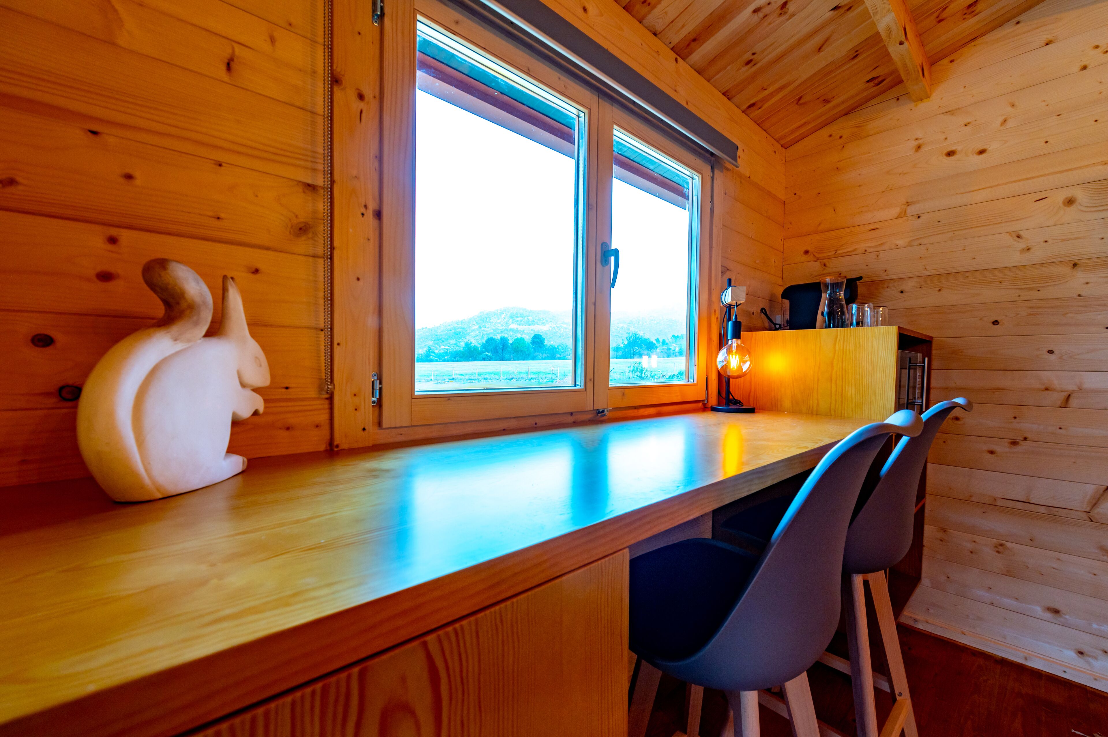 L’intérieur d’un chalet en bois, avec un comptoir en bois, deux chaises, un objet décoratif et une fenêtre donnant sur un paysage enneigé.