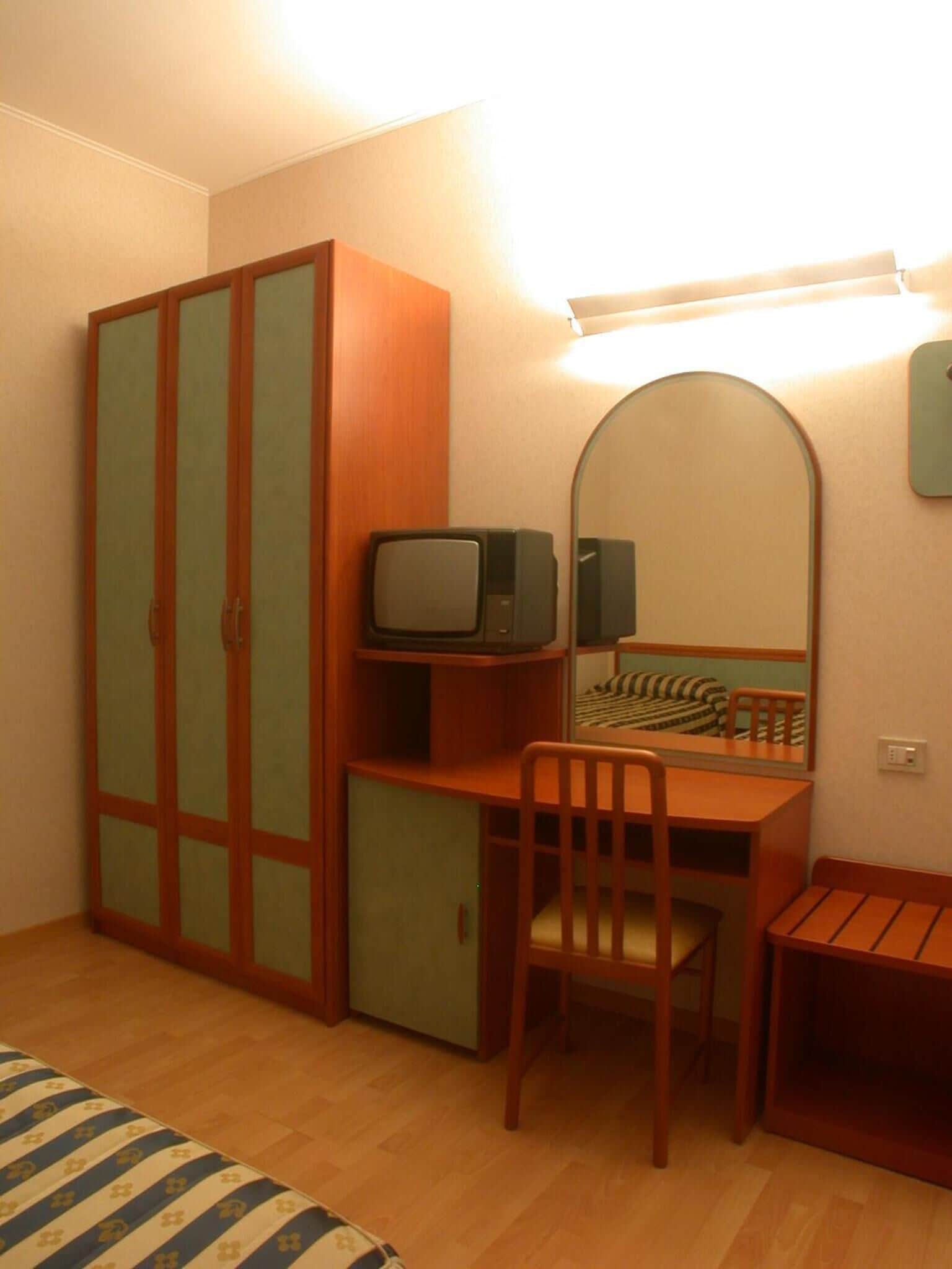 A compact bedroom with a wooden wardrobe, a small desk with a chair, a television, and a mirror.