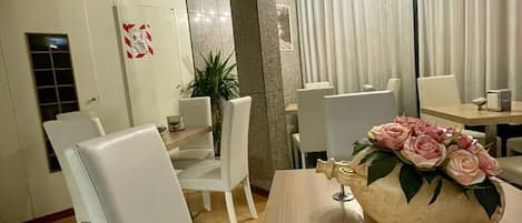 Et spiseområde med et bord dækket til to personer, hvide stole, en vase med blomster og en lysekrone.