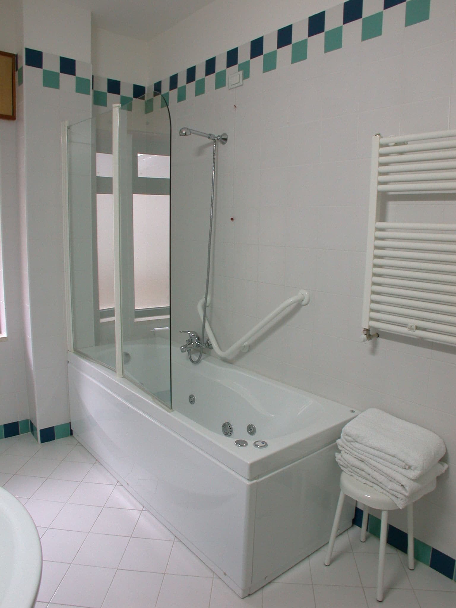 A bathroom with a glass-enclosed shower, a white bathtub, and a stack of white towels on a stool.