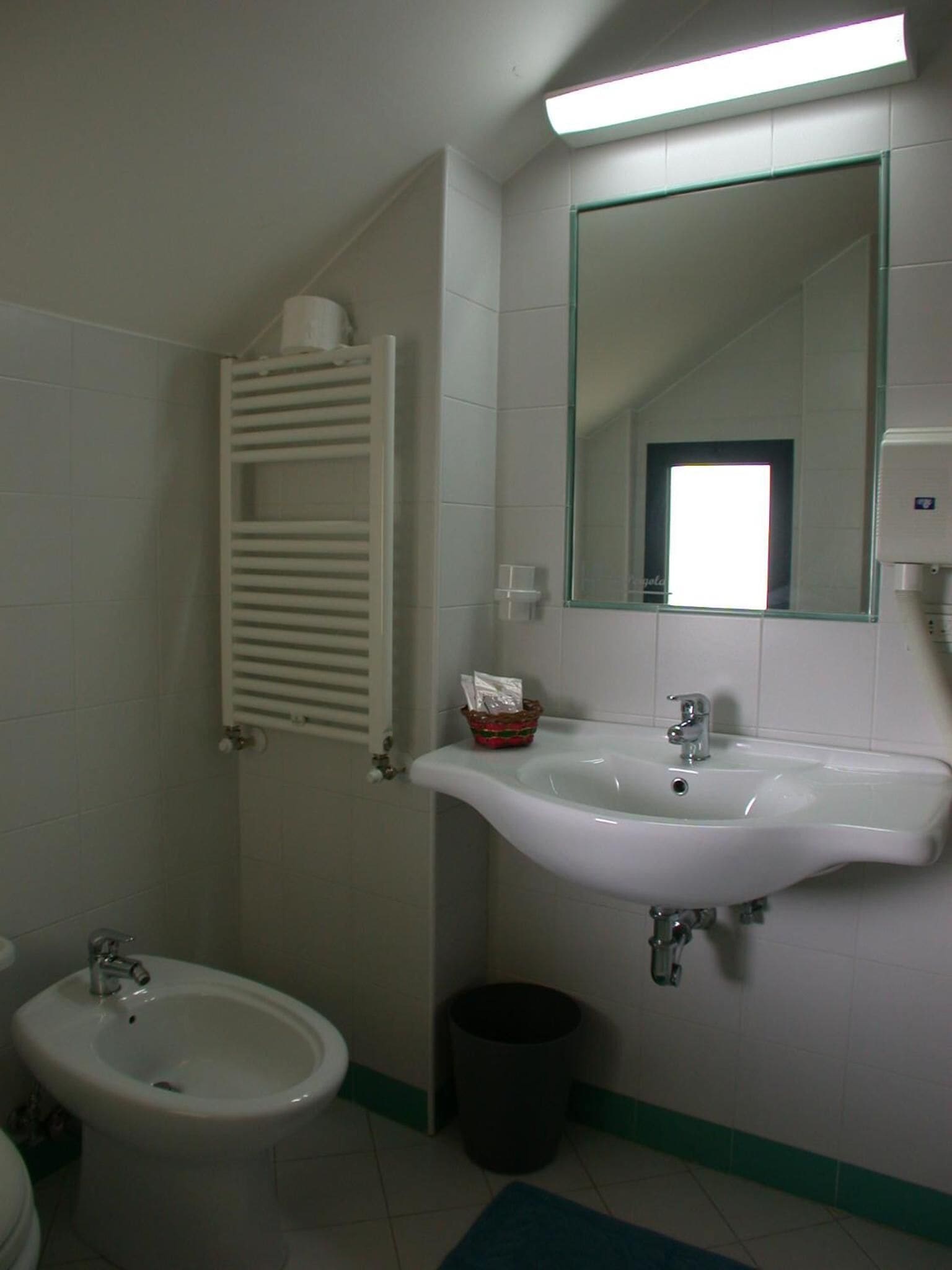 Un baño con lavamanos blanco, un bidé, toallero, basurero y espejo.