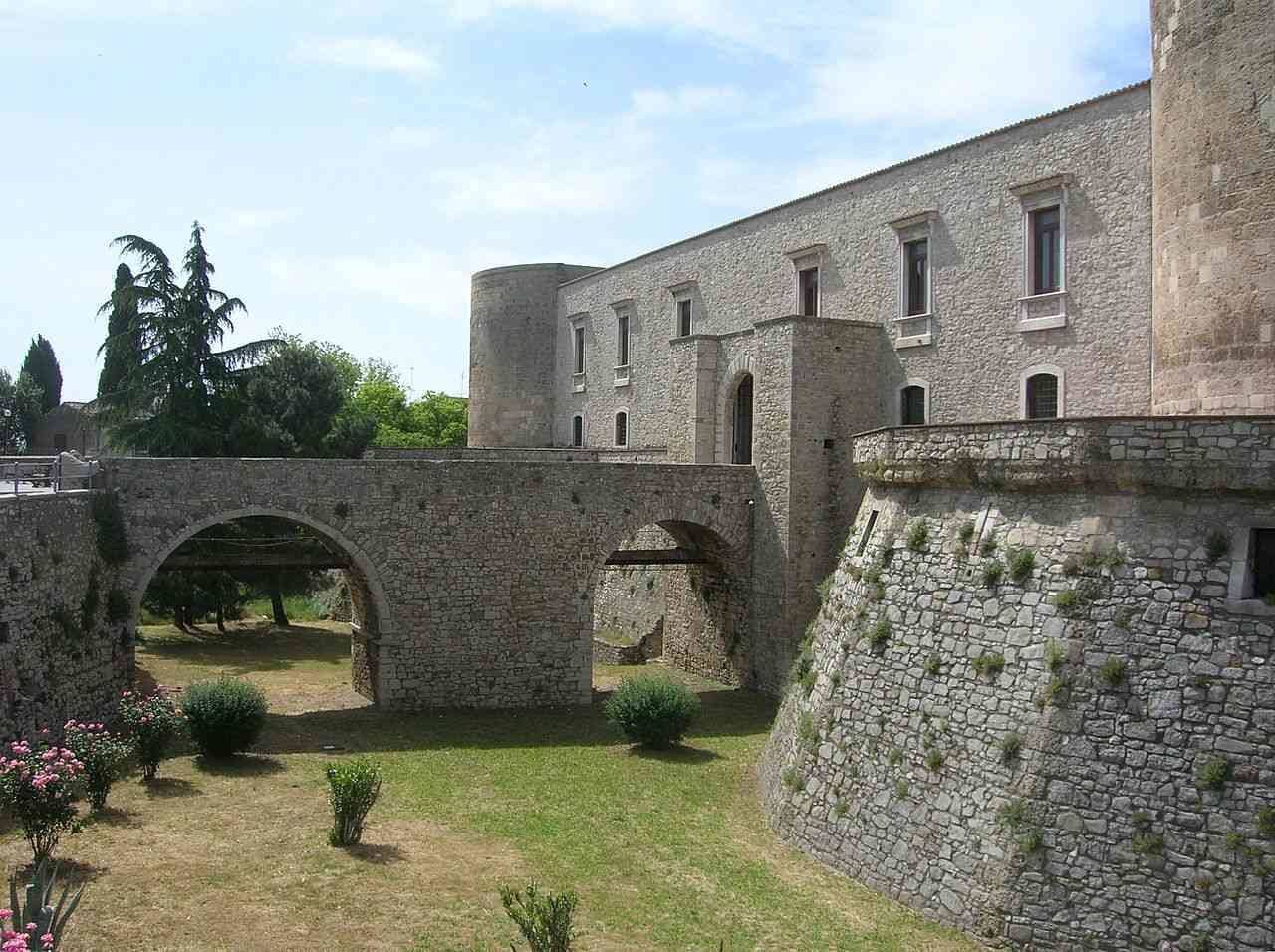 A stone building with a courtyard, an arched bridge, and a small garden with pink flowers.
