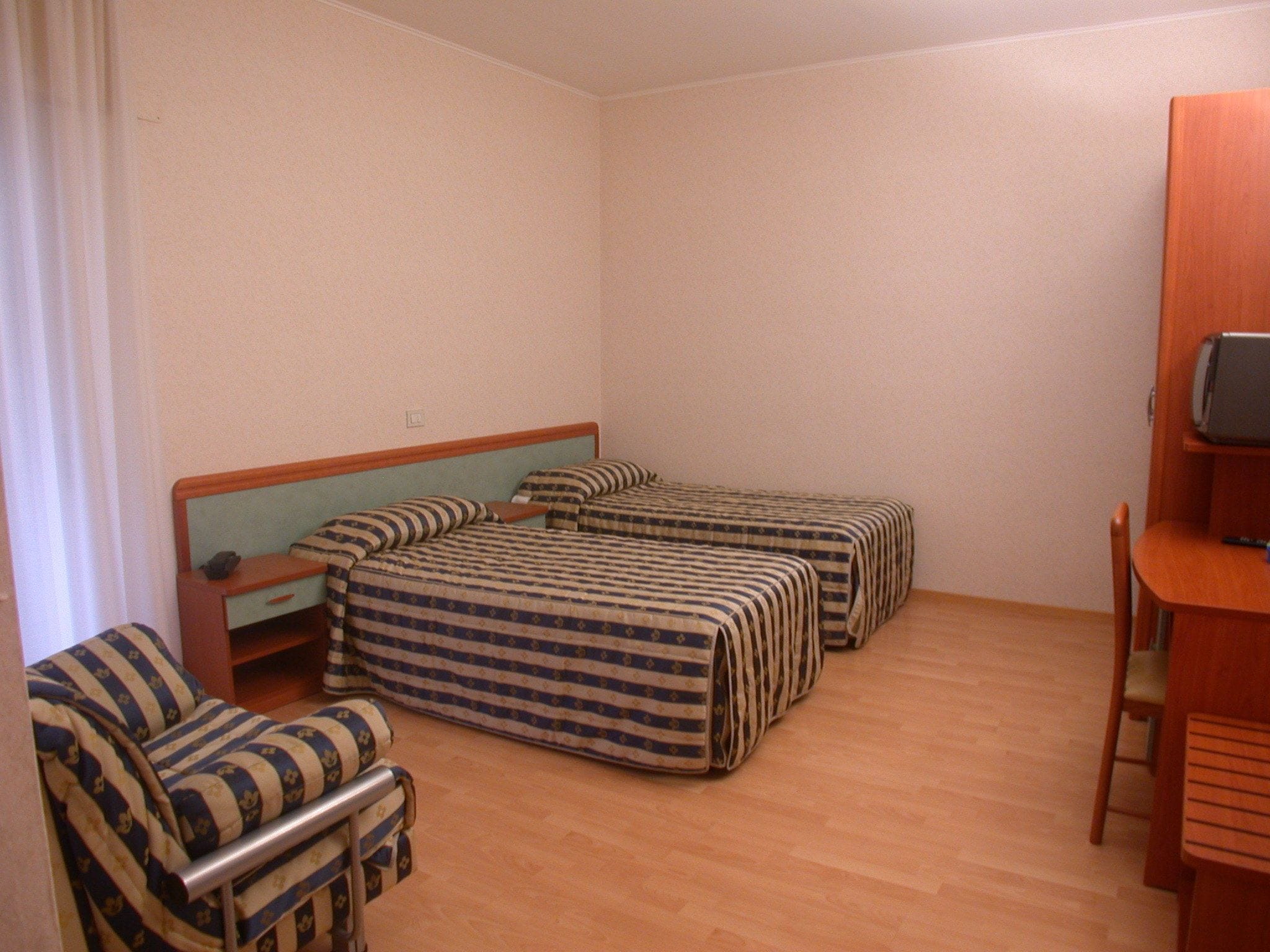 Ein Zimmer mit drei Einzelbetten, einem Holzstuhl, einem kleinen Schreibtisch mit Computer und einem Vorhang.
