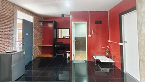 Habitación pequeña con pared de ladrillo, suelo de baldosas negras, una mesa con una silla, un cesto de basura y una puerta.