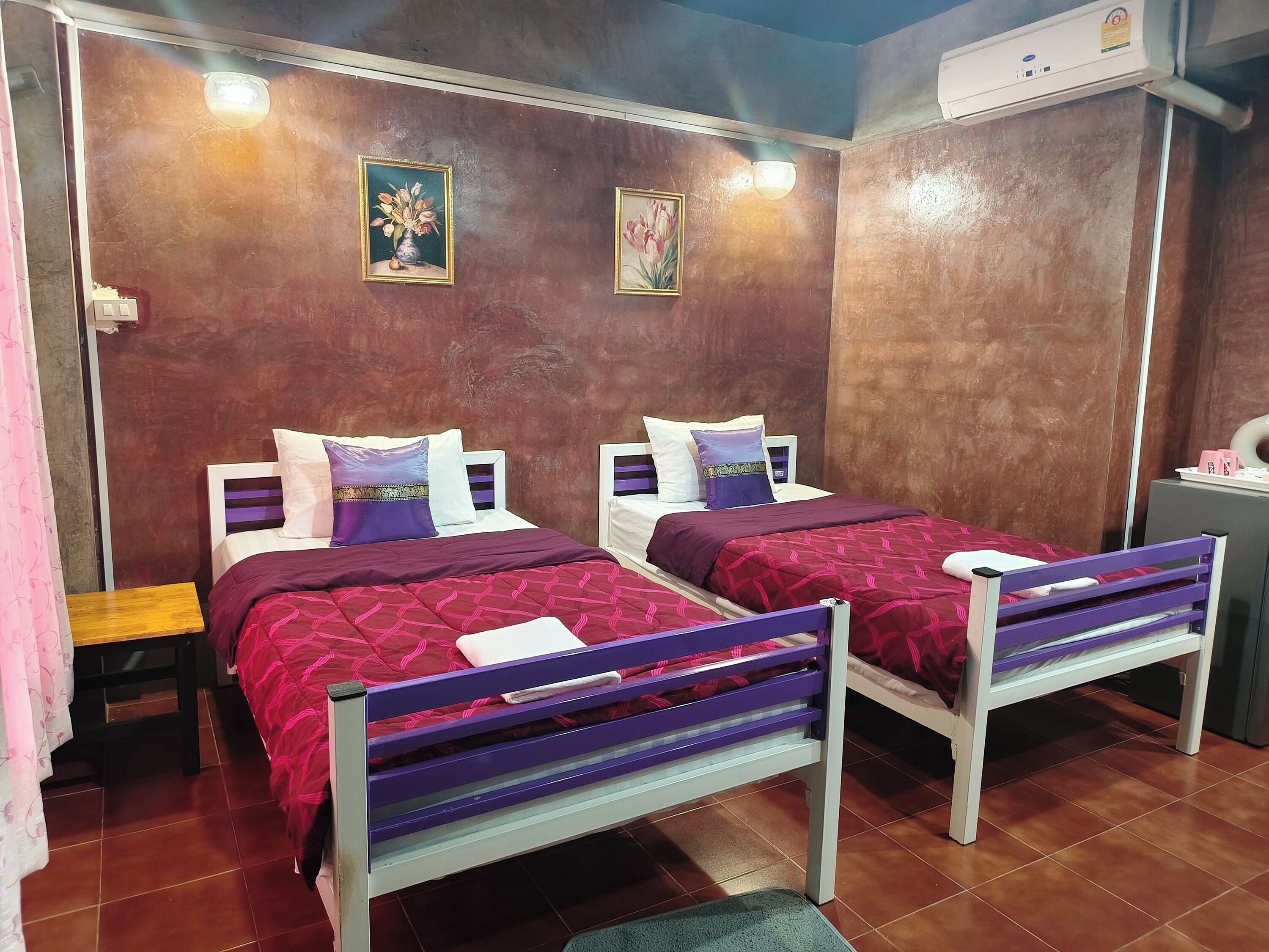 A room with two beds, each with a purple headboard and white bedding, a small wooden table, and a wall-mounted air conditioner.