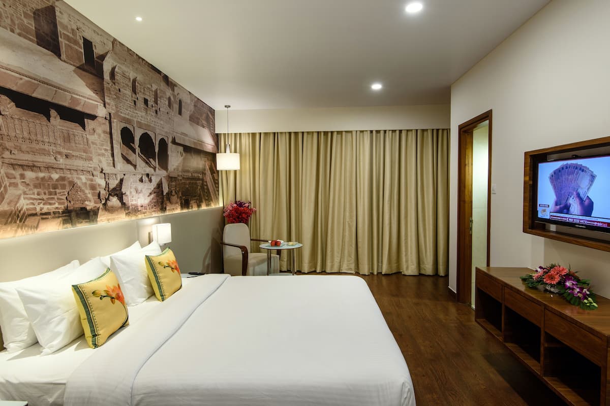 Habitación de hotel con una cama grande, un televisor de pantalla plana montado en la pared, una mesita con un jarrón de flores y una ventana cubierta por cortinas.
