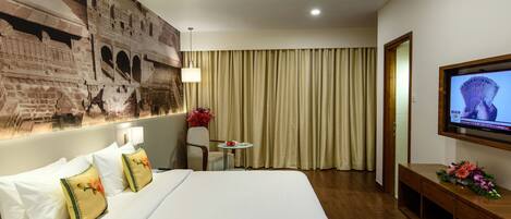 A hotel room with a large bed, a flat-screen TV mounted on the wall, a small table with a vase of flowers, and a curtain-covered window.