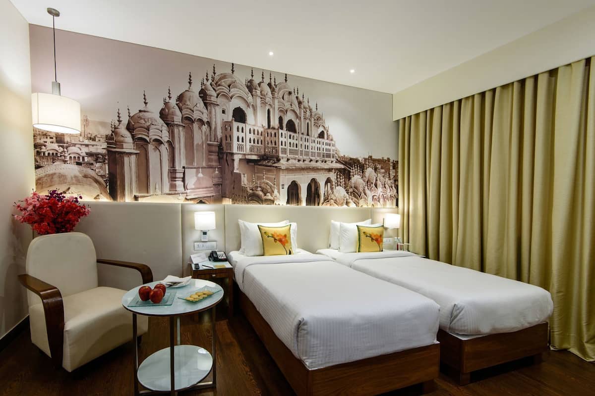 Habitación de hotel con dos camas, una silla, una mesita pequeña, una lámpara y un mural en la pared.