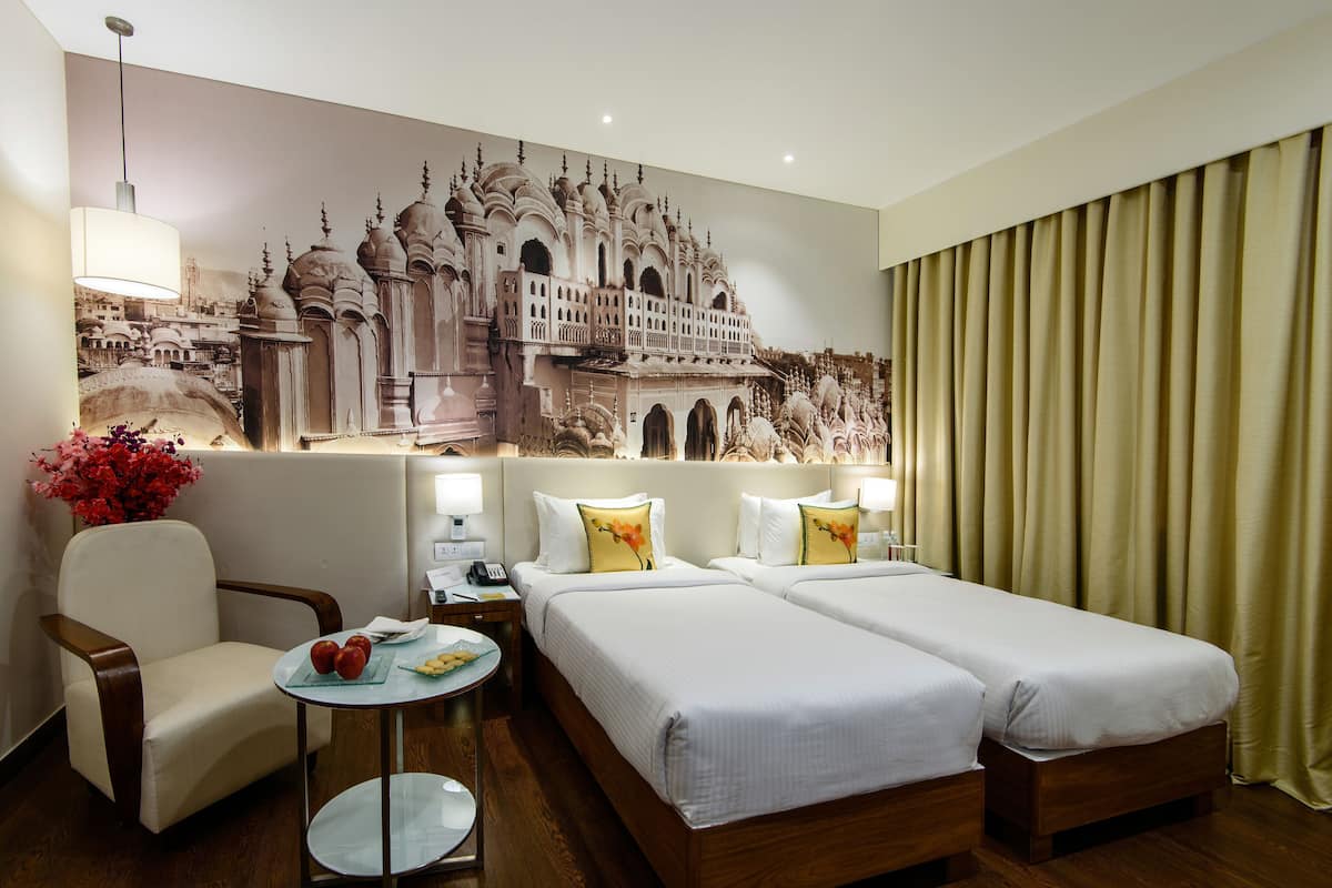 A hotel room with two beds, a chair, a small table with fruit, a lamp, and a large mural of a building.