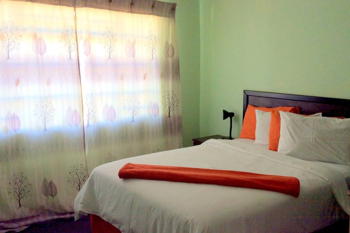 Una cama bien hecha con ropa de cama blanca y naranja, una colcha roja y un cabecero con varios almohadones.