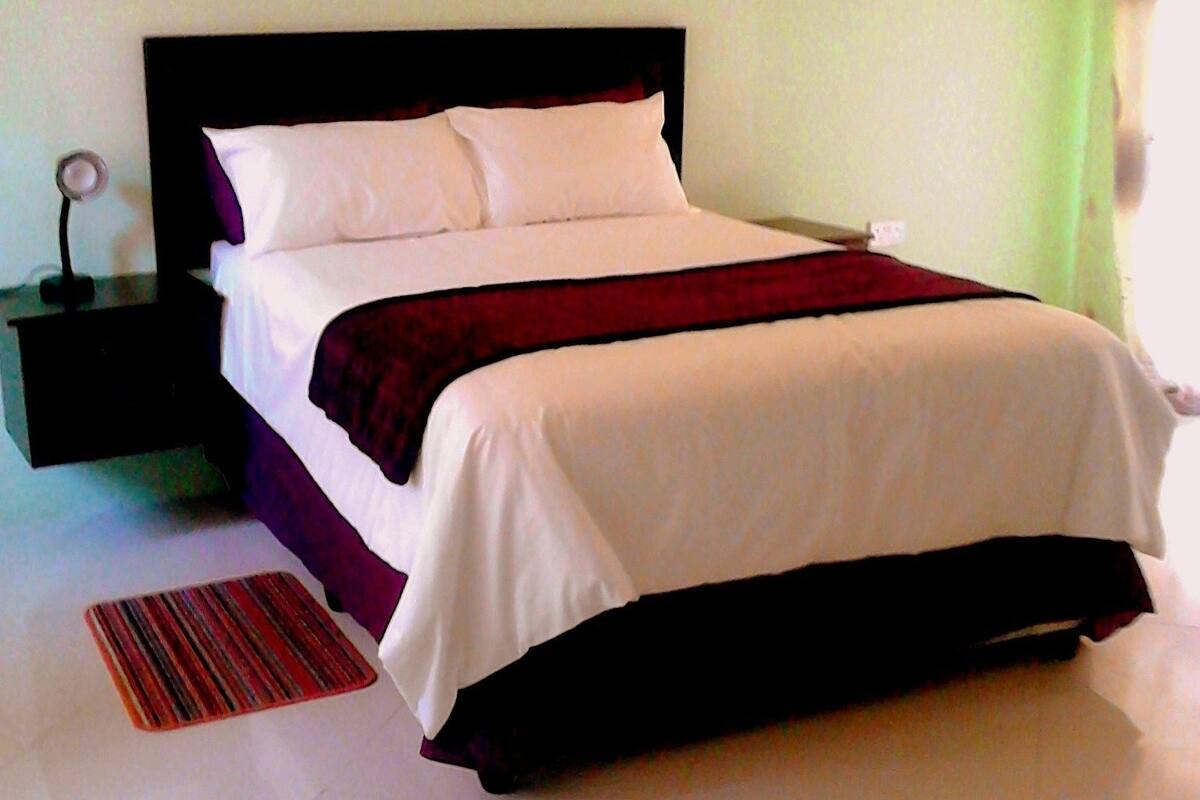 Una cama bien hecha con ropa blanca y roja, un cabecero negro, una mesita de noche con una lámpara y una alfombra a rayas rojas y negras.