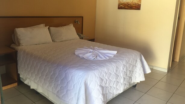 Una cama bien hecha con edredón y almohadas blancos, un cuadro de un atardecer en la pared y un piso de baldosas.