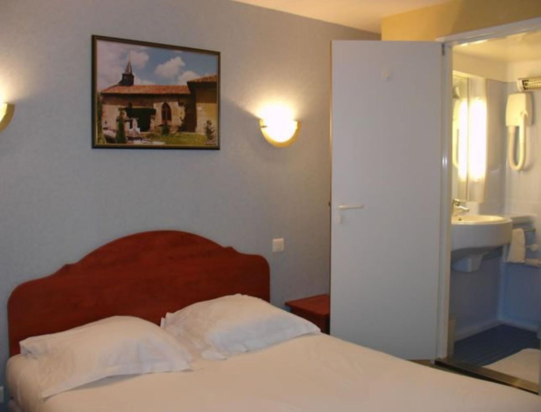 A hotel room with a bed, a painting of a building, and a bathroom visible through an open door.