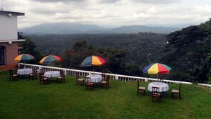 A terrace with tables and chairs, umbrellas, and a view of a mountainous landscape. - Mahaweli Regent Hotel (Kandy)