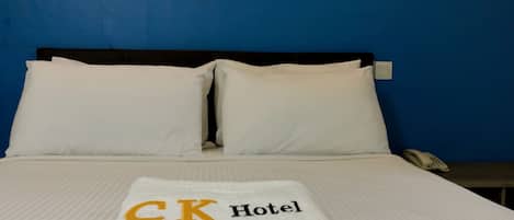 A neatly made bed with white linens and pillows, a folded white towel with "CK Hotel" embroidered on it, and a bedside telephone on a wooden nightstand.
