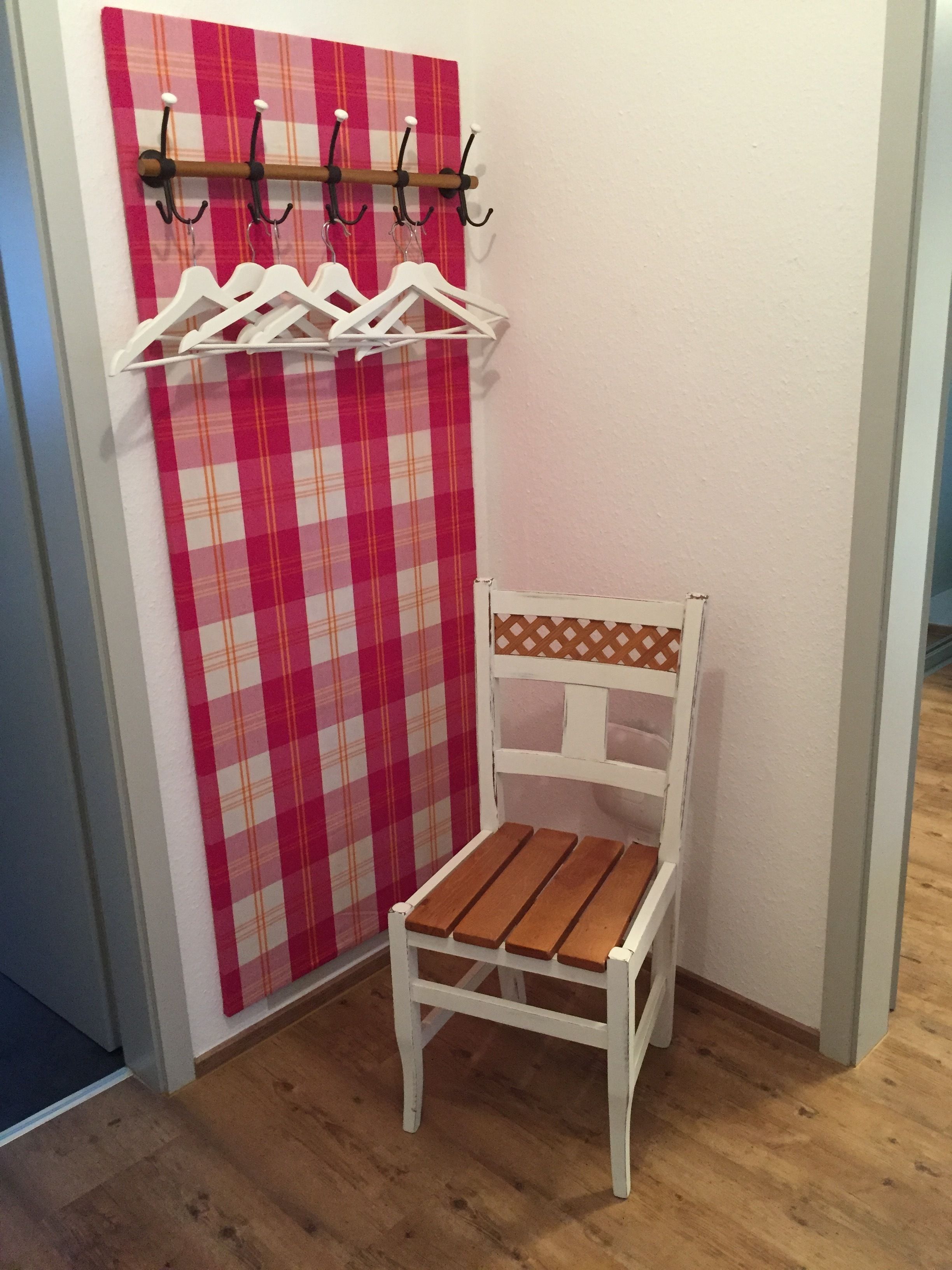 Quarto de canto com piso de madeira, uma cadeira branca com encosto estampado, cortina xadrez vermelha e branca e um porta-roupas com ganchos.