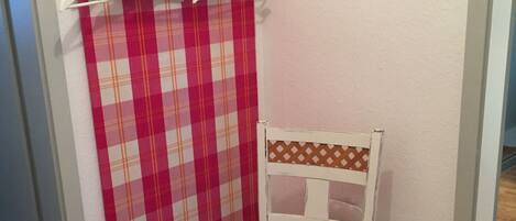 Quarto de canto com piso de madeira, uma cadeira branca com encosto estampado, cortina xadrez vermelha e branca e um porta-roupas com ganchos.