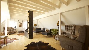 Un salon spacieux doté d’un haut plafond, de poutres en bois, d’une grande décoration représentant une tête de cerf, et de différents types de sièges, notamment un canapé, des fauteuils et une table basse.