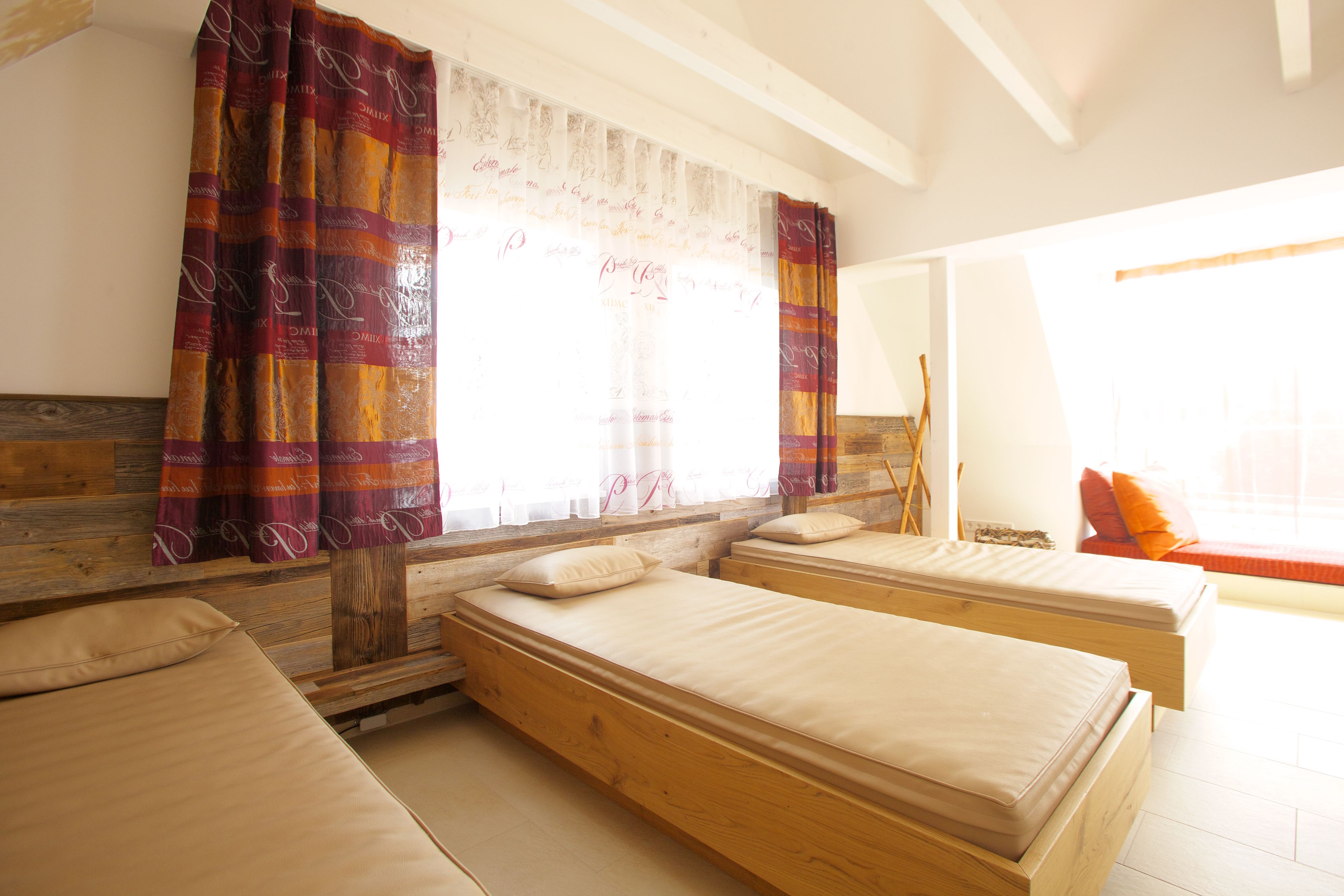 Una habitación con dos camas de madera, una ventana con cortinas, un banco de madera y un cojín rojo.