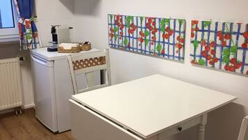 Una pequeña cocina con una mesa y una silla blancos, un refrigerador, una cafetera y un ventanal con cortinas azules.