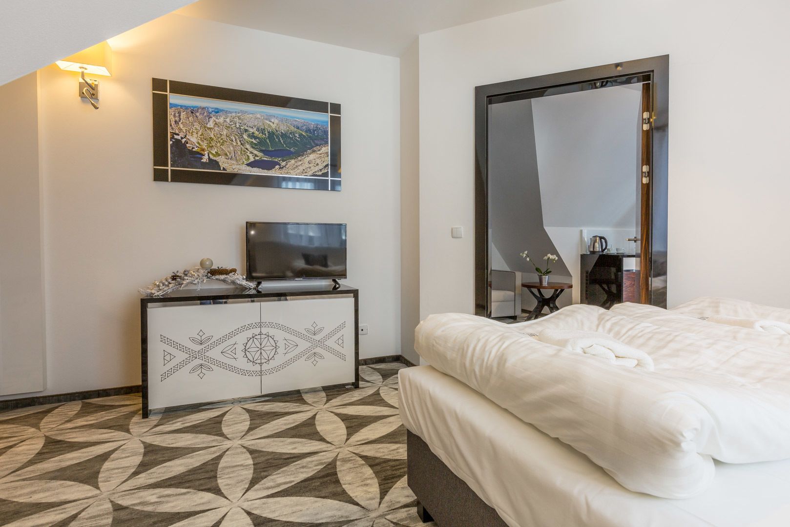 A hotel room with a bed, a TV on a white cabinet, a large mirror, and a painting of a mountainous landscape.