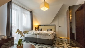 A modern hotel room with a large bed, a bedside table with a vase of flowers, a patterned carpet, and a large window with sheer curtains.