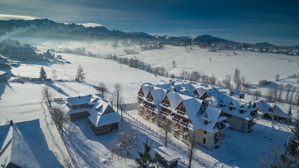 A snow-covered lodge with multiple buildings, a ski slope in the background, and mountains in the distance.