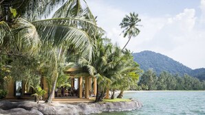 Un resort frente a la playa con un área de estar techada, palmeras y vistas al océano y las montañas.