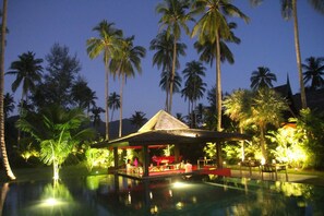 A poolside bar area with a thatched roof, surrounded by palm trees and a clear pool reflecting the night sky. - Siam Royal View Villas (Ko Chang)