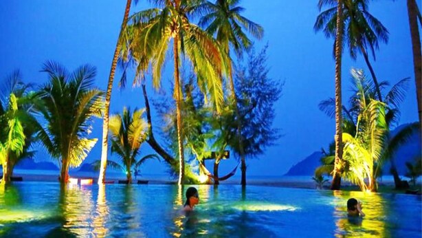 Zona de piscina con palmeras y vista al océano al atardecer.